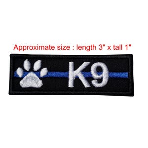 Puede incluir: Un parche rectangular negro bordado que presenta una huella de pata blanca a la izquierda y el texto K9 a la derecha, atravesado por una fina línea azul horizontal. El parche mide aproximadamente 7,62 cm de largo por 2,54 cm de alto.