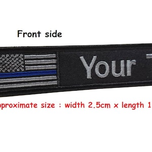 May include: Black key chain with a thin blue line American flag and the text "Your Text" embroidered in silver thread. The key chain measures 2.5 cm wide by 15.0 cm long.