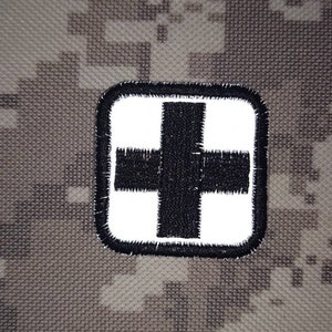 Könnte beinhalten: Weißer Stoff-Patch mit einem schwarzen Pluszeichen in der Mitte. Der Patch hat einen schwarzen Rand.