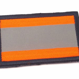 Könnte beinhalten: Ein rechteckiger schwarzer und orangefarbener Patch mit einem reflektierenden grauen Streifen in der Mitte. Der Patch hat einen schwarzen Rand.