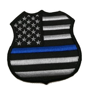 Può includere: Una toppa ricamata nera e argento a forma di distintivo della polizia con una sottile linea blu al centro. La toppa presenta una bandiera americana stilizzata con stelle bianche e strisce rosse e bianche.