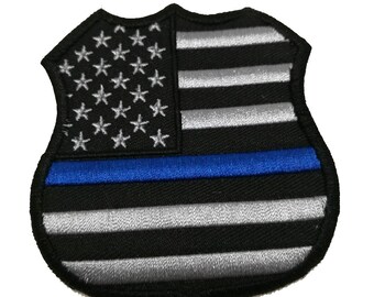 Può includere: Una toppa ricamata nera e argento a forma di distintivo della polizia con una sottile linea blu al centro. La toppa presenta una bandiera americana stilizzata con stelle bianche e strisce rosse e bianche.