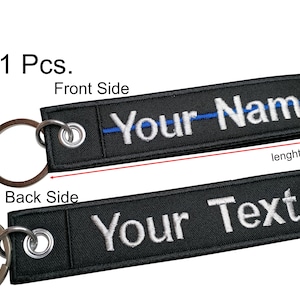 May include: Black key chain with a blue stripe and white embroidery that says "Your Name" on the front and "Your Text" on the back. The key chain is 13.5 inches long and 3 inches tall.