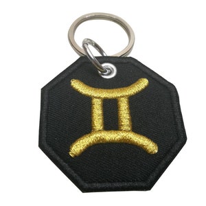 Puede incluir: Un llavero de forma octogonal negra con un símbolo zodiacal Géminis bordado en oro. El llavero tiene un anillo de metal plateado.