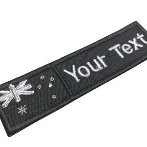 Könnte beinhalten: Ein schwarzer rechteckiger Aufnäher mit weiß gesticktem Text "Your Text". Der Aufnäher zeigt ein Flaggen-Design mit einem Stern und anderen Formen. Der Aufnäher ist wahrscheinlich für Kleidung oder Accessoires.