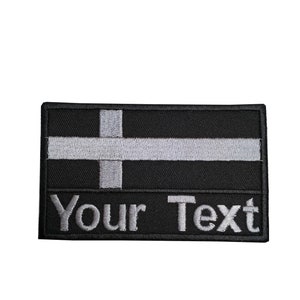 May include: Black and white embroidered patch with a white cross on a black background. The text "Your Text" is embroidered in white below the flag.