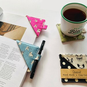 Può includere: Un set di tre segnalibri in tessuto e due sottobicchieri in tessuto con un motivo di Bulldog Francese. I segnalibri sono rosa, blu e verde. I sottobicchieri sono bianco e nero con pois e beige con un motivo di Bulldog Francese. I sottobicchieri sono etichettati "Original Book Mark x Cup Coaster 9 cm x 9 cm | Cotton & Linen blend".