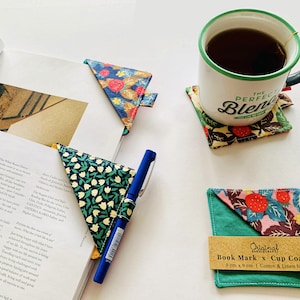 Può includere: Un set di due segnalibri e sottobicchieri in tessuto fatti a mano. I segnalibri sono triangolari e presentano un motivo floreale in tonalità di blu, verde e giallo. I sottobicchieri sono quadrati e presentano un motivo floreale in tonalità di rosa, verde e marrone. I sottobicchieri hanno un retro in tessuto verde. Il testo "Original Book Mark x Cup Coaster 9 cm x 9 cm | Cotton & Linen blend" è stampato su un'etichetta bianca attaccata al sottobicchiere.
