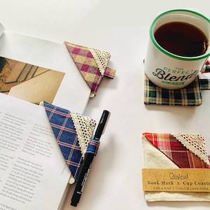 Può includere: Una collezione di segnalibri fatti a mano e un sottobicchiere. I segnalibri presentano tessuto a quadri con finiture in pizzo. Una penna è appoggiata su un libro con un segnalibro a quadri blu. Una tazza di tè è su un sottobicchiere a quadri. Le dimensioni sono 9 cm x 9 cm.