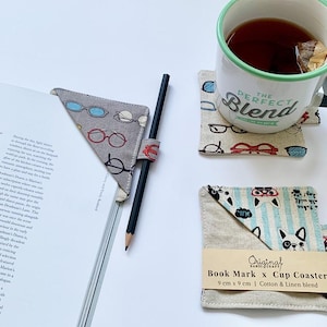 Può includere: Una tazza di tè poggia su un sottobicchiere con occhiali. Un segnalibro con occhiali è su un libro con una matita. È presente anche un secondo set di sottobicchieri e segnalibri. La tazza dice "THE PERFECT BLEND".