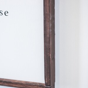 May include: Close-up of a framed sign with a dark brown wooden frame. The sign has white space with black text. The frame is made of dark brown wood and is mounted on a white wall.