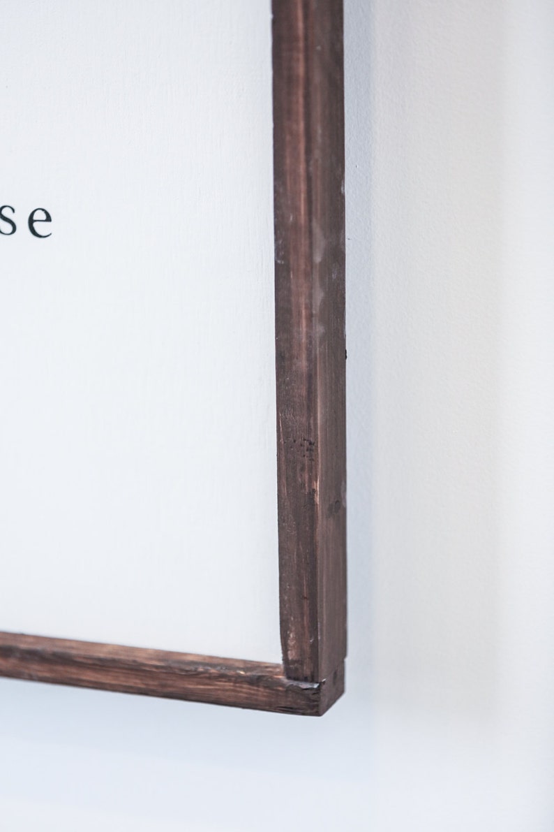 May include: Close-up of a framed sign with a dark brown wooden frame. The sign has white space with black text. The frame is made of dark brown wood and is mounted on a white wall.