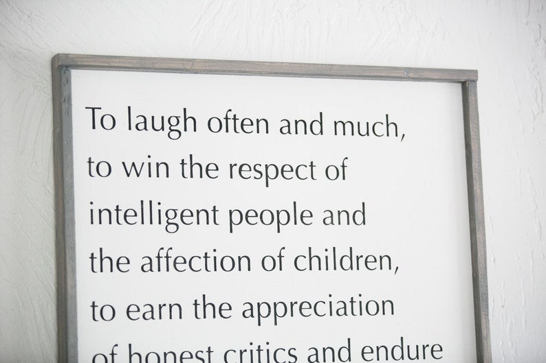 May include: A framed white sign with black text that reads "To laugh often and much, to win the respect of intelligent people and the affection of children, to earn the appreciation of honest critics and endure..." The frame is a muted gray color.