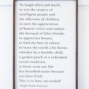May include: A framed white sign with black text quoting Ralph Waldo Emerson: "To laugh often and much... This is to have succeeded!" The frame is a dark brown color. The sign is a decorative piece.