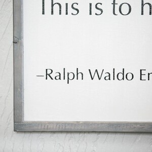 May include: A framed white sign with black text that reads "This is to have..." followed by "-Ralph Waldo Emerson." The frame is a weathered gray color. The background is a textured white wall.