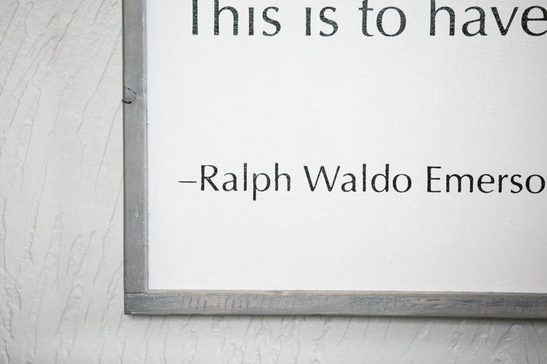 May include: A framed white sign with black text that reads "This is to have..." followed by "-Ralph Waldo Emerson." The frame is a weathered gray color. The background is a textured white wall.