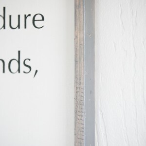 May include: A close-up of a wooden framed sign with text. The frame is a combination of gray paint and natural wood. The sign is on a white wall. The text on the sign is partially visible.