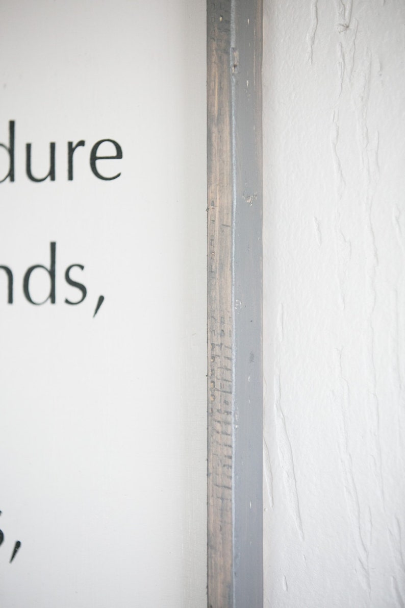 May include: A close-up of a wooden framed sign with text. The frame is a combination of gray paint and natural wood. The sign is on a white wall. The text on the sign is partially visible.