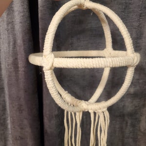 May include: Cream-colored macrame plant hanger with a spherical design. The hanger features a circular base and a series of intersecting arches, all crafted from thick, woven rope. Long, dangling fringe hangs from the bottom, adding a bohemian touch.