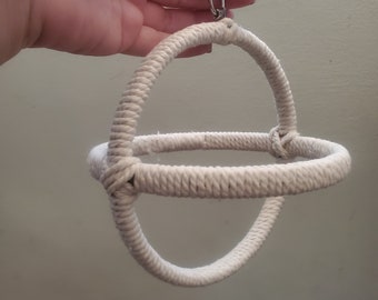 May include: A handmade, off-white rope toy for pets, featuring a circular design with a connecting horizontal loop. A silver metal carabiner clip is attached for hanging. The rope is tightly wound, creating a textured surface.