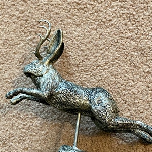 May include: A silver-toned sculpture of a jackalope, a mythical creature with a hare's body and deer antlers, in mid-jump. The sculpture is mounted on a round, distressed base and supported by a thin metal rod. The base is approximately 10 cm in diameter.