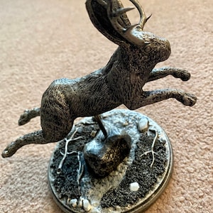 May include: A silver-toned sculpture of a hare with antlers, in mid-leap. The sculpture sits on a circular base with a textured, snow-like design. The hare's fur is detailed, and the antlers are a striking feature.