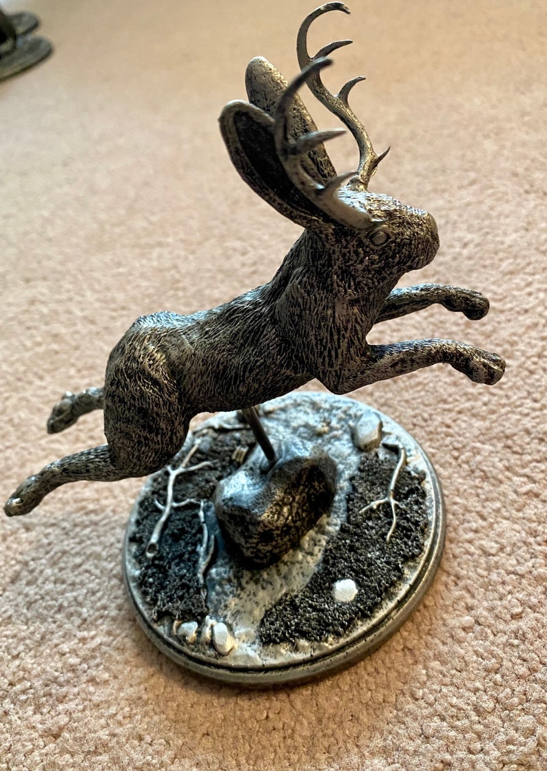May include: A silver-toned sculpture of a hare with antlers, in mid-leap. The sculpture sits on a circular base with a textured, snow-like design. The hare's fur is detailed, and the antlers are a striking feature.