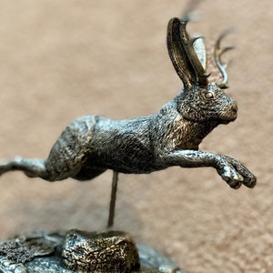 May include: A silver-toned sculpture of a hare in mid-leap, featuring antlers. The hare has detailed fur and is mounted on a textured base. This decorative sculpture would make a great gift.