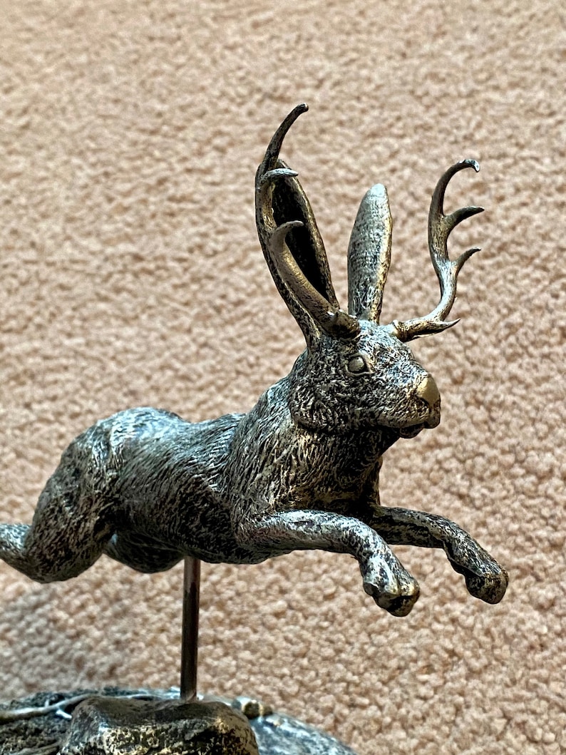 May include: A silver-coloured sculpture of a hare with antlers, captured mid-jump. The sculpture has a textured finish and is mounted on a metal rod fixed to a stone-like base. The hare's ears are erect, and its antlers are prominent.