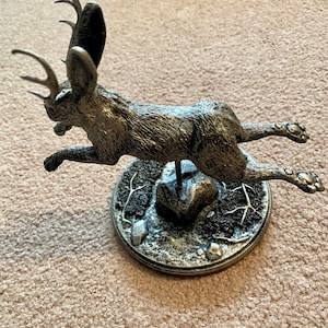 May include: A silver jackalope sculpture in mid-leap. The mythical creature has antlers and large ears. It is mounted on a circular base with a textured, rocky appearance. The sculpture is detailed and appears to be made of metal.