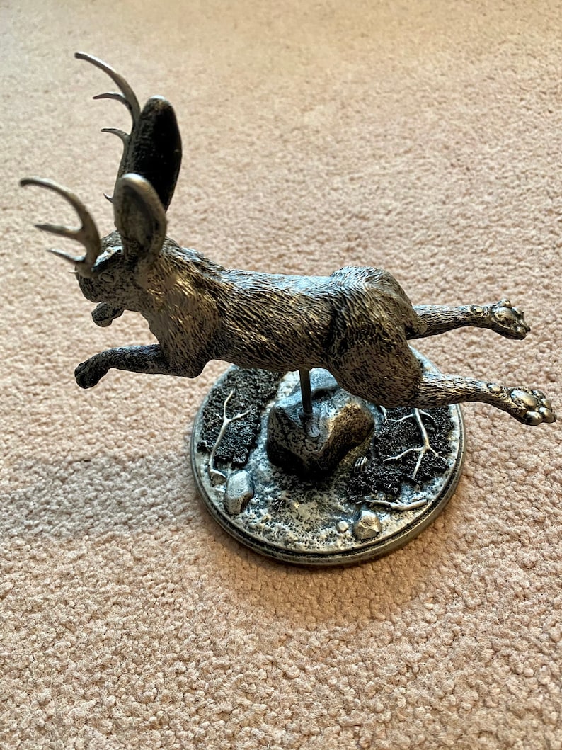 May include: A silver jackalope sculpture in mid-leap. The mythical creature has antlers and large ears. It is mounted on a circular base with a textured, rocky appearance. The sculpture is detailed and appears to be made of metal.