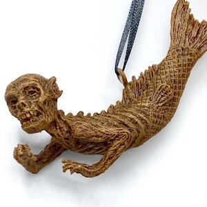 May include: A brown, skeletal mermaid ornament with a detailed, textured surface. The ornament is hanging from a black ribbon.