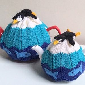 Könnte beinhalten: Zwei gestrickte Teekannenwärmer mit Pinguinen darauf. Die Wärmer sind blau, weiß und lila mit einem Fischdesign.