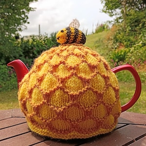 May include: A yellow and brown knitted tea cozy with a honeycomb pattern, topped with a small bee. The tea pot has a red handle and spout. The cozy is designed to keep tea warm and adds a decorative touch to tea time.
