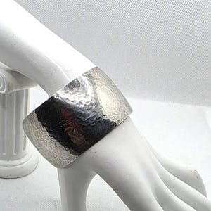 May include: A wide, silver-toned cuff bracelet featuring a textured, hammered surface. The metal band has a polished finish that reflects light across its curved, rigid structure, creating a series of small, irregular indentations.