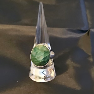 May include: A silver ring featuring a large, faceted, emerald-green gemstone. The ring is displayed on a clear, triangular stand against a dark background. The gemstone has a round shape and a brilliant cut.