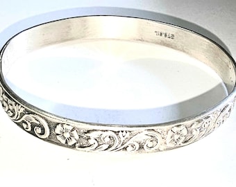May include: A silver-toned bangle bracelet with an engraved floral and scroll design. The bracelet has a smooth, polished finish on the inside and a detailed pattern on the outside. The word "STEEL" is stamped on the inside of the bracelet.