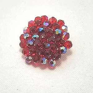 May include: A round, red beaded brooch. The brooch is composed of many small, faceted beads in varying shades of red, with some beads displaying iridescent color. The beads are clustered together to form a circular shape.