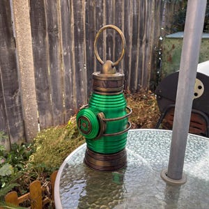 May include: A cylindrical green lantern with a metallic bronze finish on the top and bottom sections. The lantern features a prominent circular emblem on the side, horizontal ribbed detailing along the body, and a large circular handle at the top.