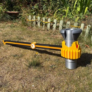 Puede incluir: Un martillo de utilería grande con un mango largo negro con una franja amarilla central y un emblema circular. La cabeza del martillo es plateada con un diseño cilíndrico acanalado y una base amarilla brillante con un pequeño diamante rojo.