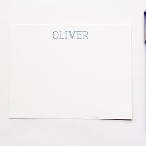 May include: A white stationery set with the name "OLIVER" printed in blue. A blue pen is positioned next to the stationery. The pen is a vibrant blue color. The stationery is rectangular and has a simple, elegant design.