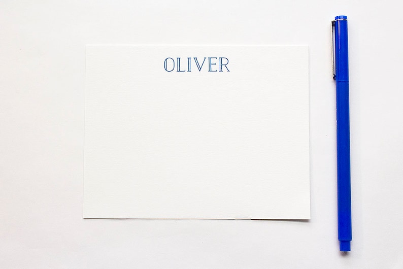 May include: A white stationery set with the name "OLIVER" printed in blue. A blue pen is positioned next to the stationery. The pen is a vibrant blue color. The stationery is rectangular and has a simple, elegant design.