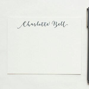 May include: A white stationery card with the name "Charlotte Bell" in cursive script. A gray pen is positioned vertically to the right of the card. The background is a plain white surface, creating a clean and minimalist aesthetic.