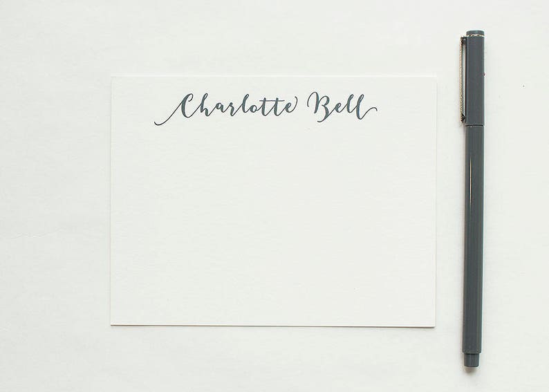 May include: A white stationery card with the name "Charlotte Bell" in cursive script. A gray pen is positioned vertically to the right of the card. The background is a plain white surface, creating a clean and minimalist aesthetic.