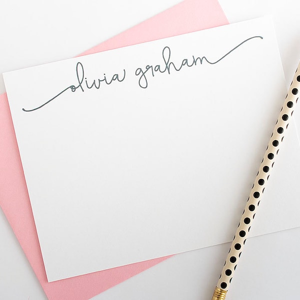 May include: A white stationery set featuring a personalized notepad with the name "olivia graham" in cursive script, resting on a pink envelope. A pencil with black polka dots and a gold eraser is placed diagonally.