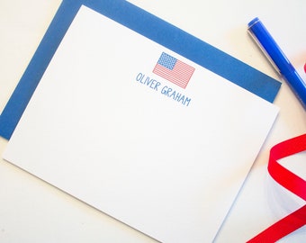 Könnte beinhalten: Weißes Briefpapier mit blauem Umschlag, mit einer gedruckten amerikanischen Flagge und dem Namen "OLIVER GRAHAM" in Blau. Ein blauer Stift und ein rotes Band sind ebenfalls vorhanden, was ein patriotisches Thema schafft.