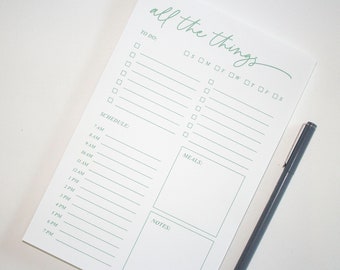 Op de afbeelding: Een witte notitieblok met de woorden "all the things" in groen schrift. Het notitieblok heeft secties voor takenlijsten, een schema, maaltijden en notities. Een donkergrijze pen ligt aan de rechterkant van het notitieblok.