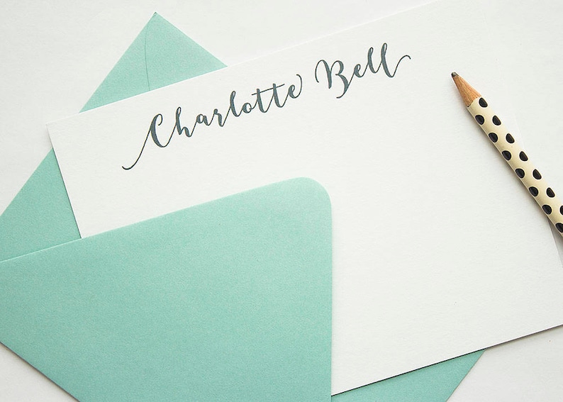 May include: A stationery set featuring a white card with the name "Charlotte Bell" in elegant script, nestled in a light teal envelope. A pencil with a black polka dot design rests beside the card, suggesting a writing theme.