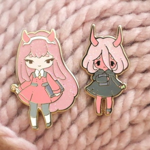 May include: Two enamel pins featuring anime-style characters with pink hair and horns. One pin depicts a character in a pink dress and the other in a gray dress. Both pins have gold-colored metal borders and are holding books.