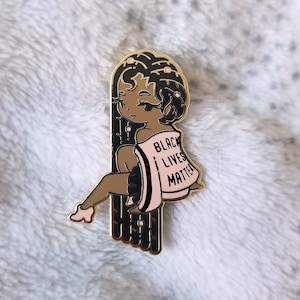 May include: Gold enamel pin featuring a cartoon illustration of a Black woman with long braids, wearing a pink shirt with the text "Black Lives Matter" printed on it.